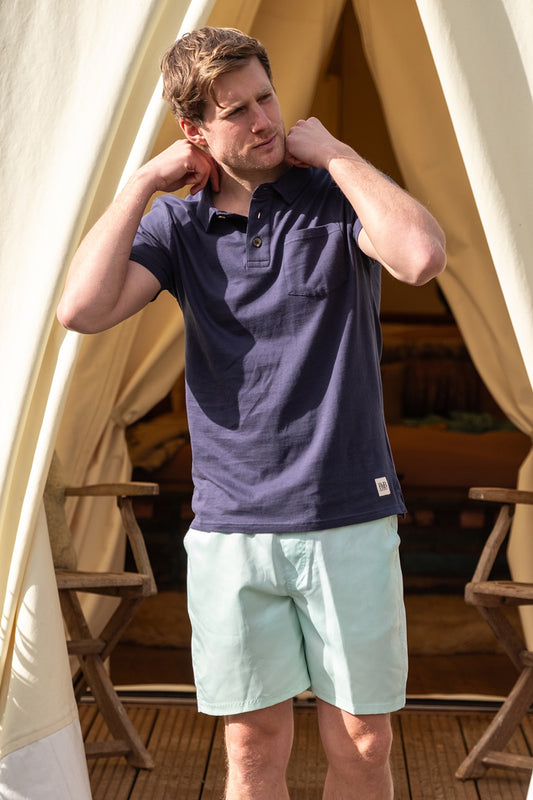 Lifestyle image of male model standing in front of a tipi tent wearing a blue polo shirt and azure coloured swim shorts