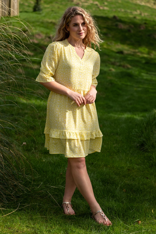 Sunny lifestyle image of a female model wearing a yellow ruffled dress from Rupert and Buckley stood on some grass