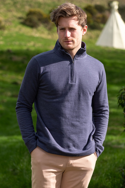 Lifestyle image of male model wearing a blue 1/4 jumper stood against a grass field and tent in the background